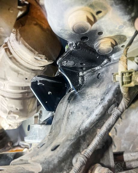 Black FMI front differential mount bracket welded to the driver side frame rail of a Ford Bronco, viewed from underneath