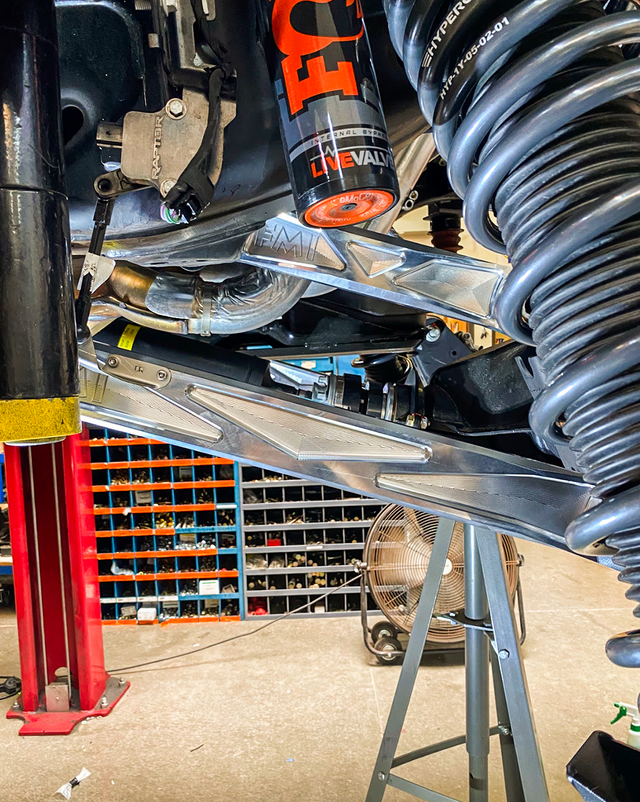 Raw billet aluminum rear links installed on a Ford Bronco Raptor on the shop lift, next to the factory FOX rear shock